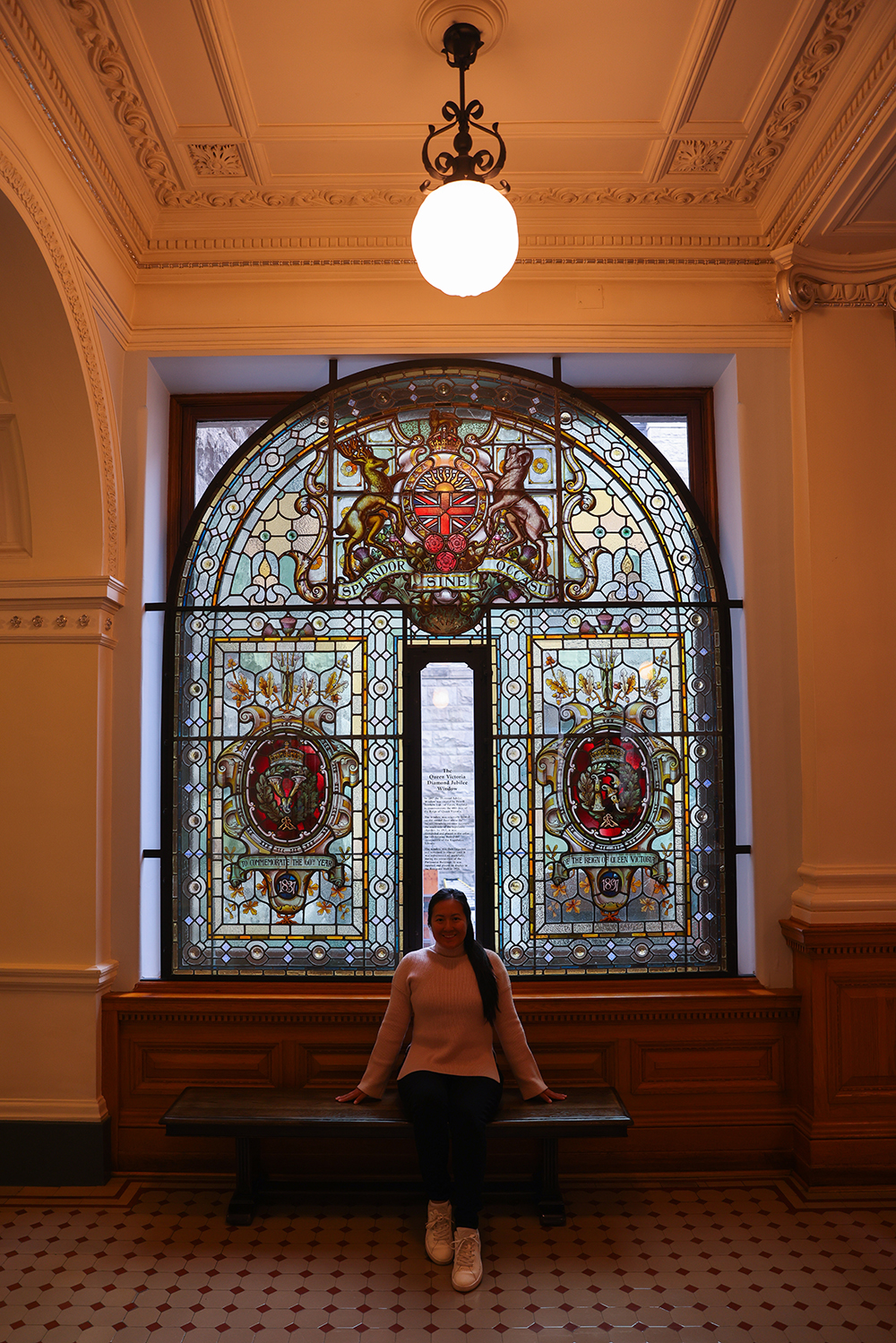Asian woman wears a light pink sweater, black jeans, and white shoes by a huge glass window with mosaic arts inside the British Columbia Parliament buildings