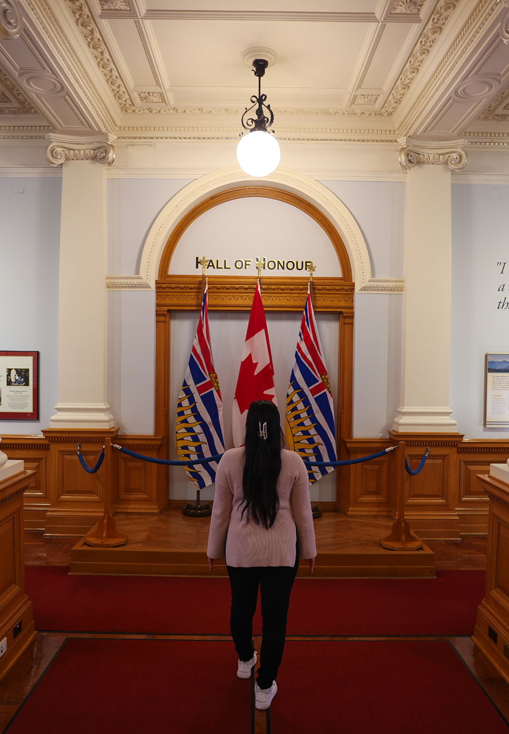 Asian woman wears a light pink sweater, black jeans, and white shoes by Canadian flag and BC flags inside the British Columbia Parliament buildings