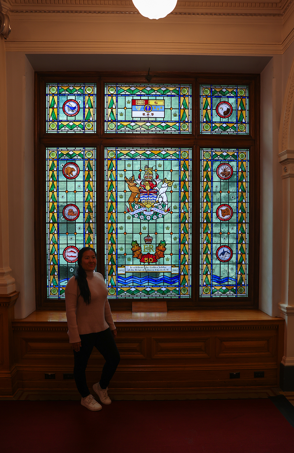 Asian woman wears a light pink sweater, black jeans, and white shoes by a huge glass window with mosaic arts inside the British Columbia Parliament buildings