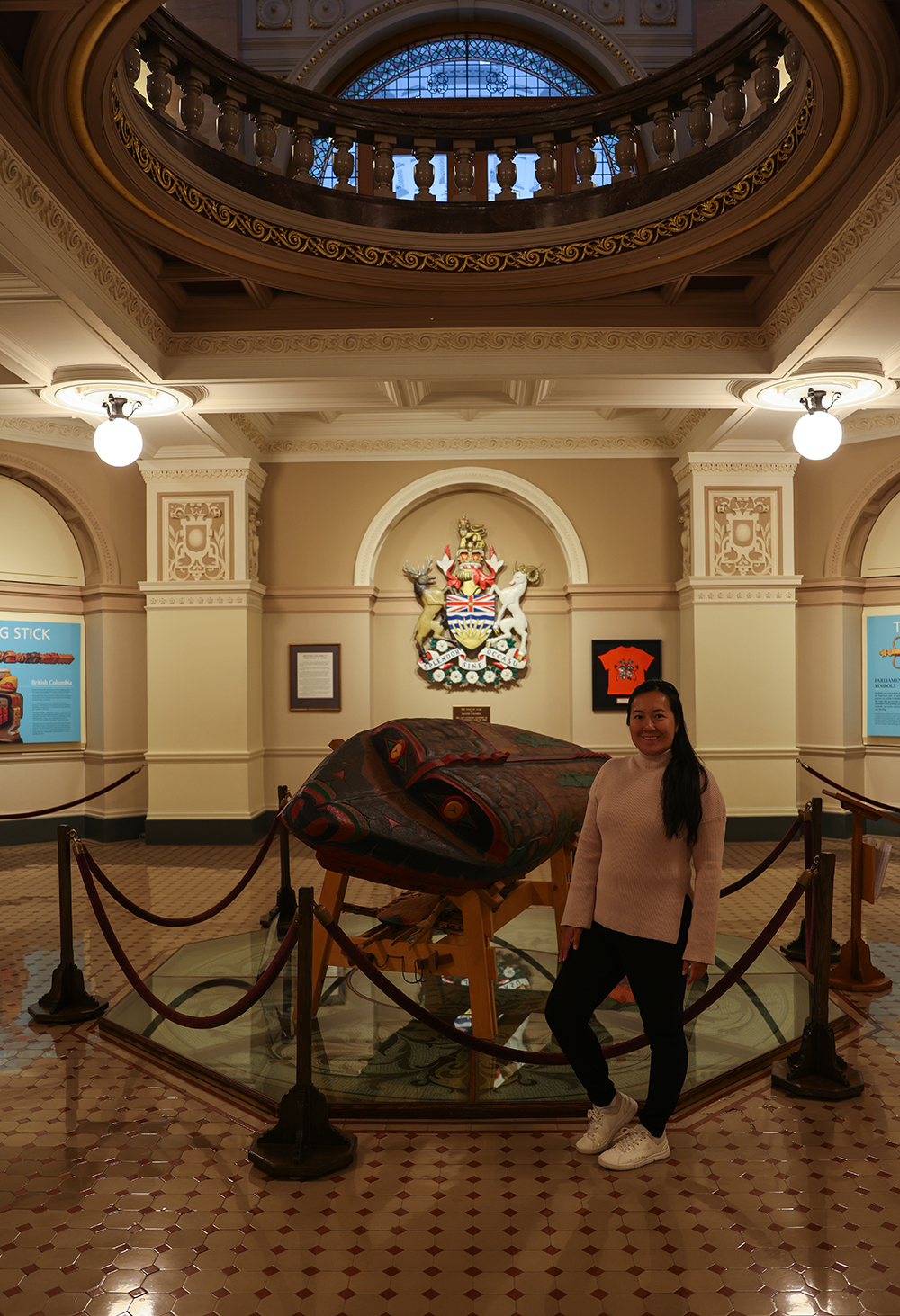 Asian woman wears a light pink sweater, black jeans, and white shoes by an native art sculpture inside the British Columbia Parliament buildings