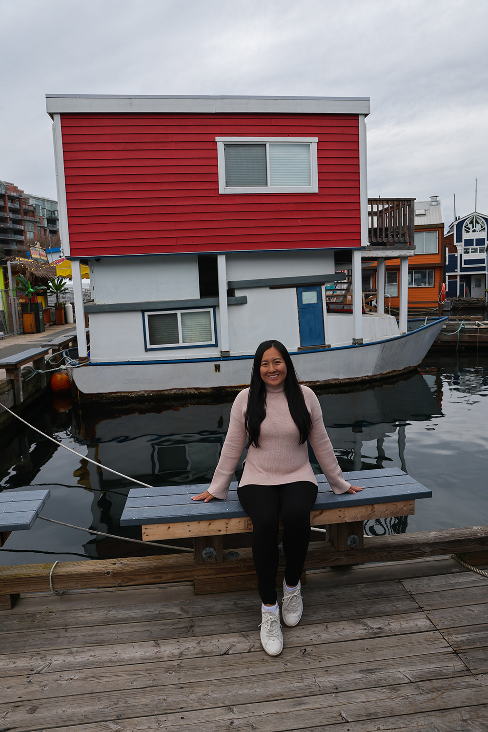 Asian woman wears a light pink sweater, black jeans, and white shoes sitting on the bench at the Fisherman's Wharf, Victoria, British Columbia BC