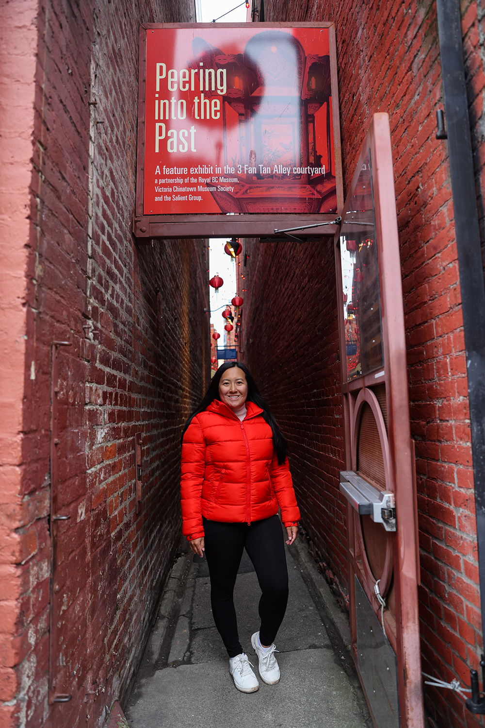 Asian woman wears a red jacket, black jeans, and white casual shoes, standing in the front of Fan Tan Alley entrance in Chinatown, Victoria, British Columbia Canada