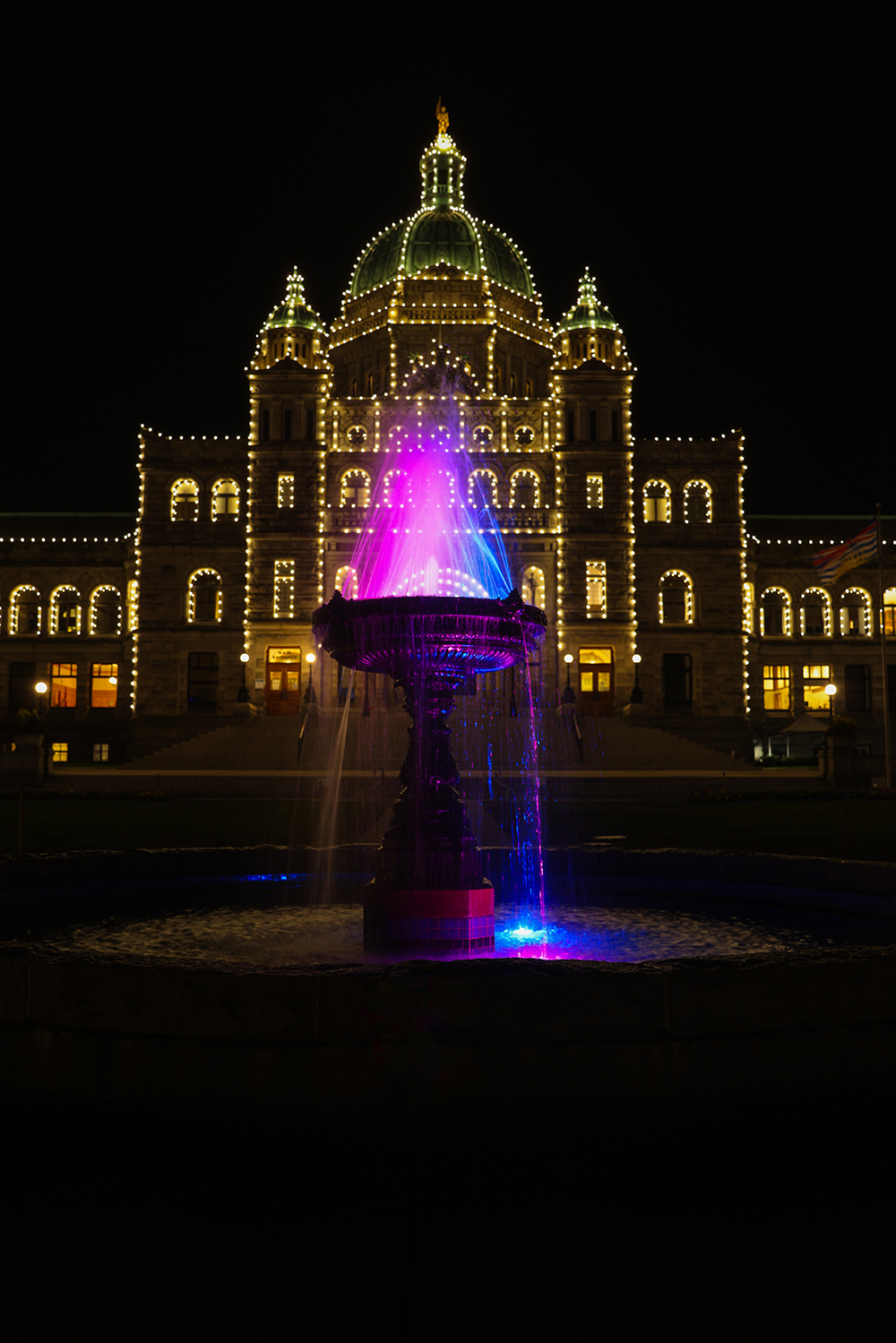 British Columbia Parliament Buildings with yellow lights on and water fountain with blue and pink lights in Victoria, BC Canada