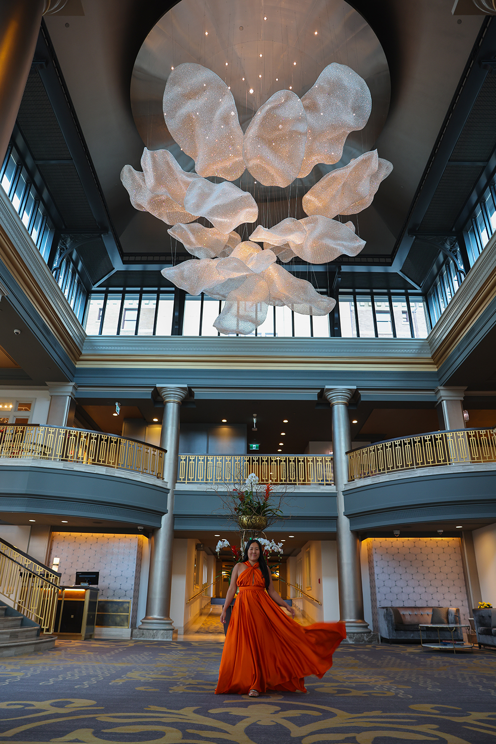 Asian woman wears a flowy orange dress inside the front entrance of the Fairmont Empress hotel with a huge chandelier in Victoria, BC