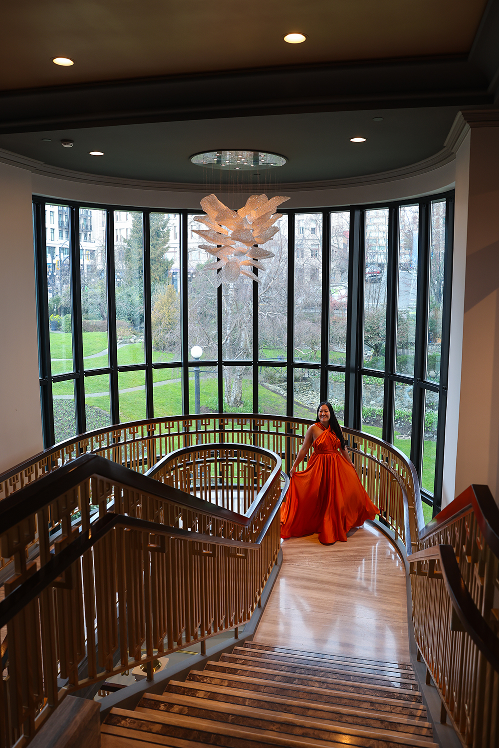 Asian woman wearing an flowy orange dress on the staircase by the beautiful, huge glass windows with a small light chandelier inside the Fairmont Empress hotel, Victoria, BC