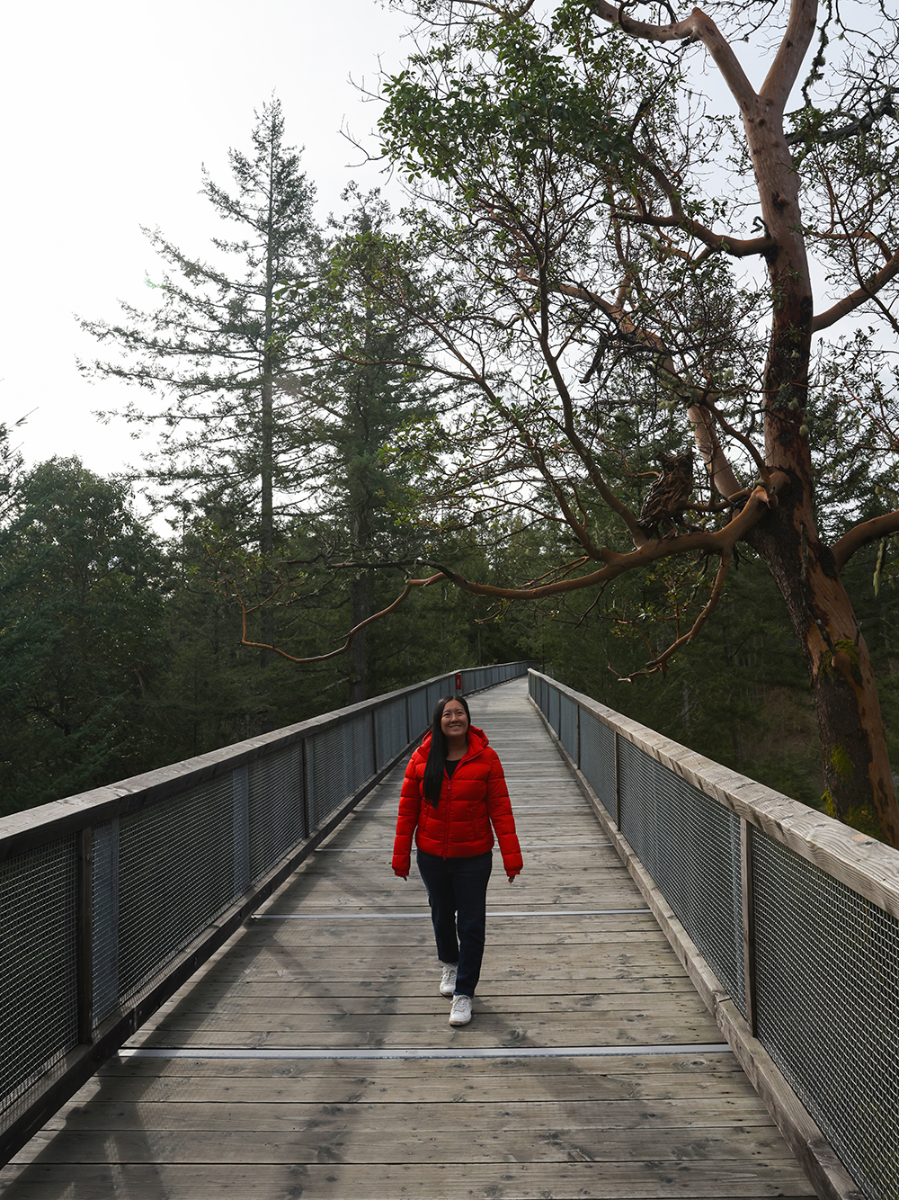 Asian woman wears a red jacket, blue jeans, and white casual shoes walking on a walkway through the forest by a tree with a wooden shape of the owl at the Malahat SkyWalk