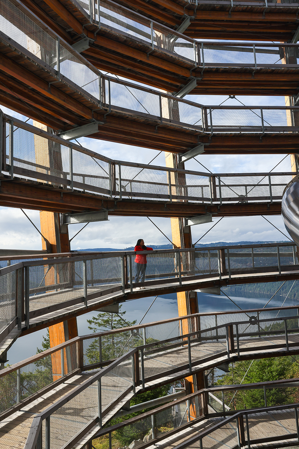 Asian woman wears a red jacket, blue jeans, and white casual shoes standing and crossing her arms on the railing at the Spiral Tower of the Malahat Skywalk