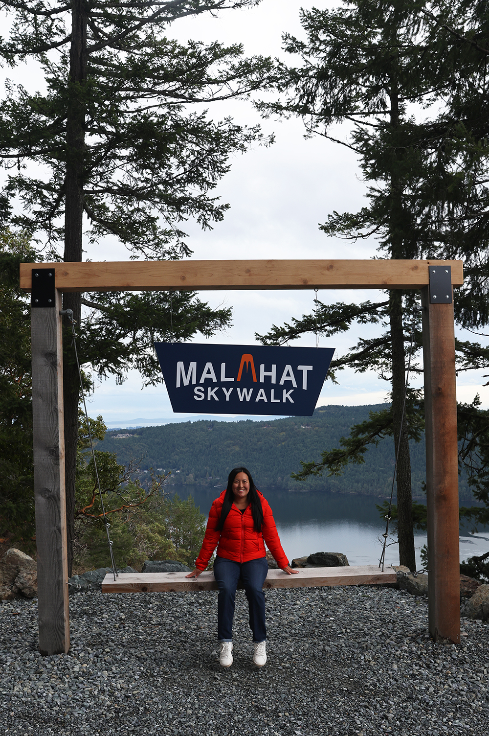 Asian woman wears a red jacket, blue jeans, and white casual shoes on a giant wooden swing with a sign of Malahat Skywalk by the trees in the background of the Saachet Inlet