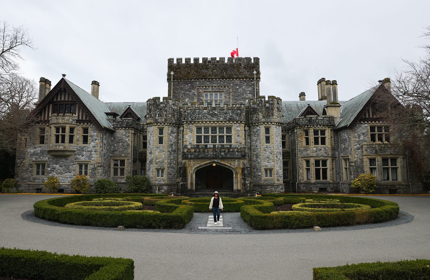 Asian woman wear black shirt, white sweater with flowers, blue jeans, and white shoes, and tan hat in the front of the Hatley Castle, Victoria, BC Canada
