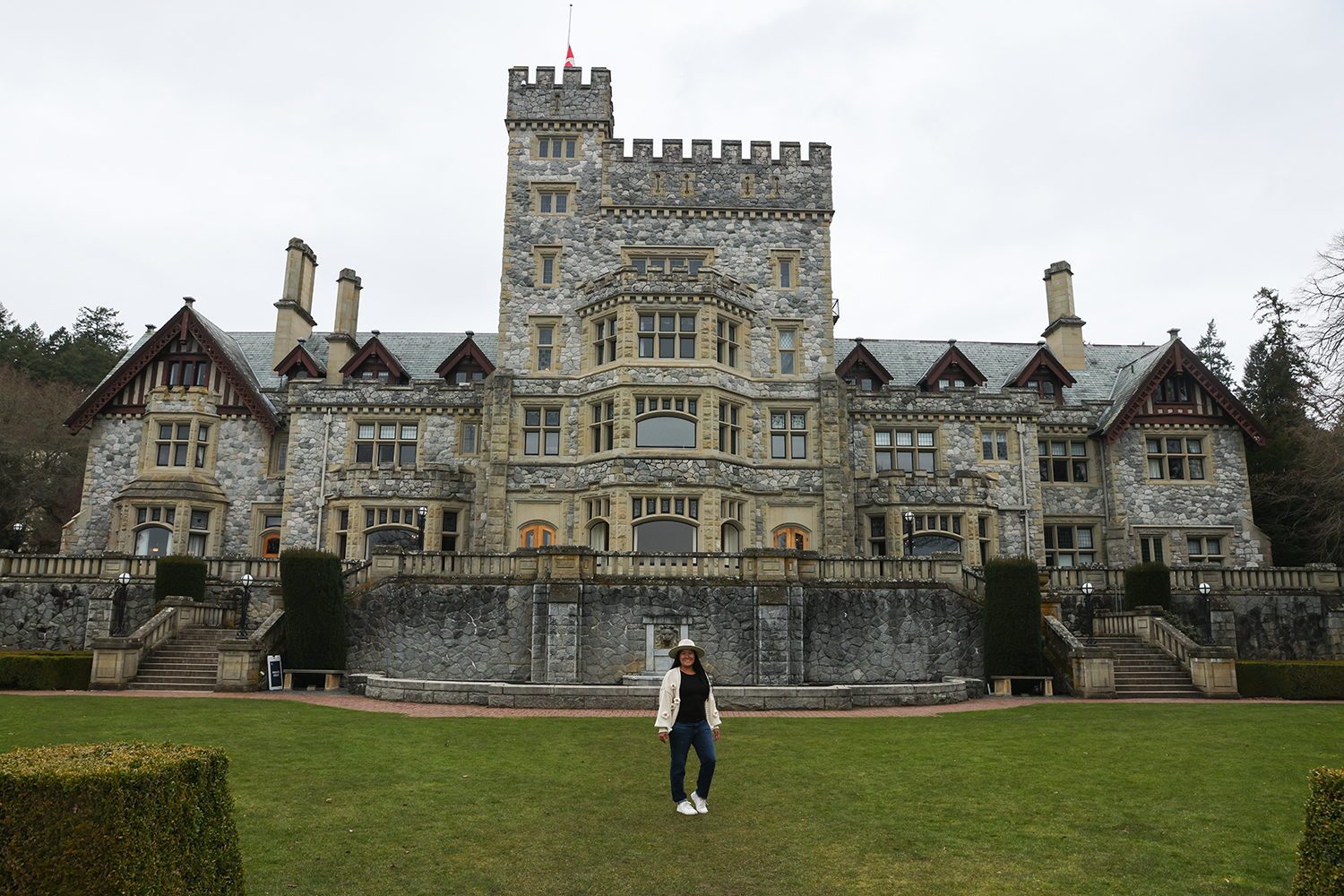Asian woman wear black shirt, white sweater with flowers, blue jeans, and white shoes, and tan hat in the back of the Hatley Castle, Victoria, BC Canada