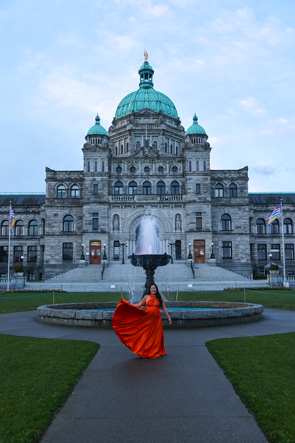 Asian woman wears a flowy orange dress in the front of British Columbia Parliament Buildings in Victoria