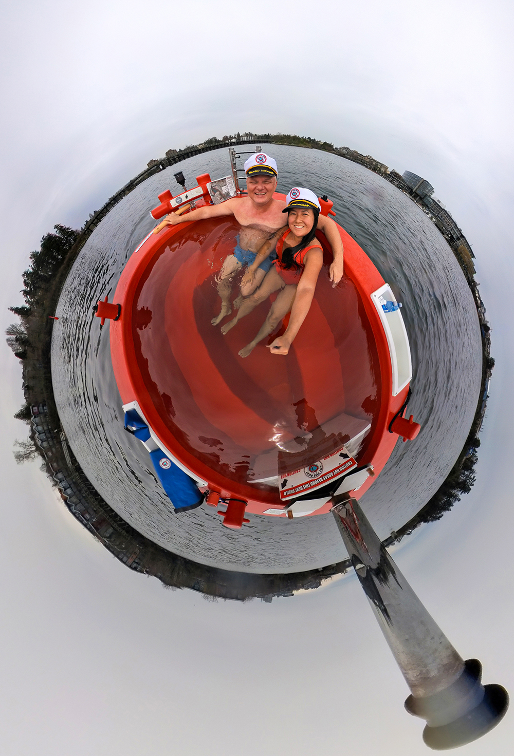 A white man and an Asian woman, both wearing swimsuits, relax in a hot tub boat as they cruise through Victoria, BC's picturesque Inner Harbour.
