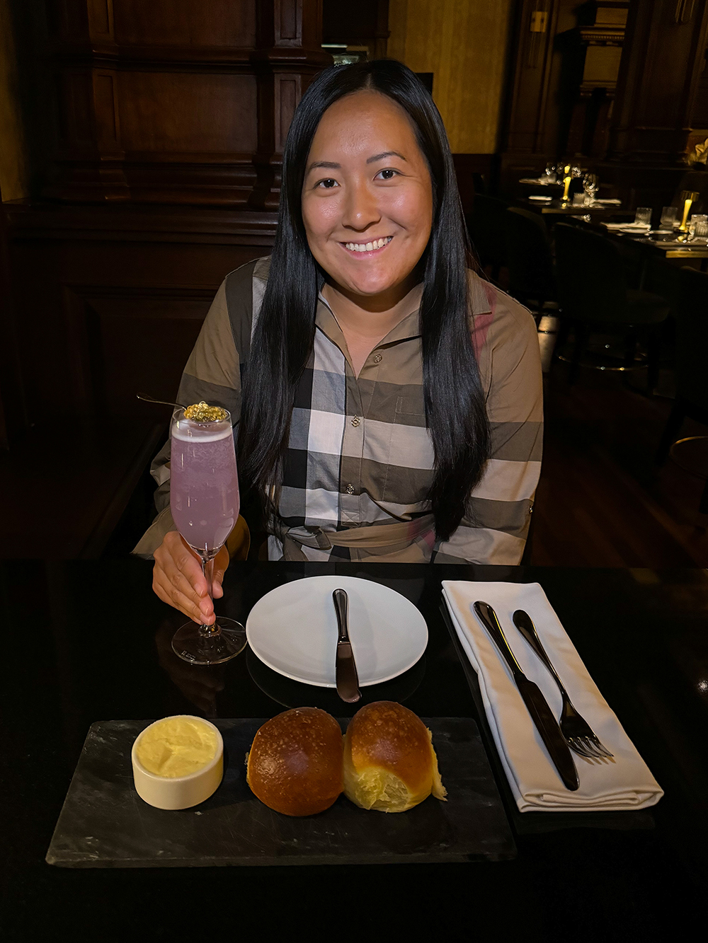 An Asian woman wears Burberry plaid dress, sits and holds a Empress Cocktail with soft breads and butter at the Q at the Fairmont Empress.