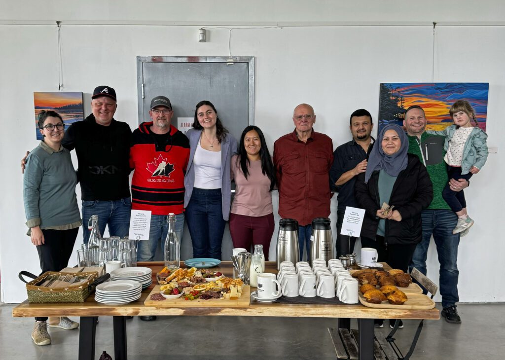 10 people stand and pose happily together by a wooden table with coffee, charcuterie boards, and muffins at the Fantastico bar-deli, Victoria, BC