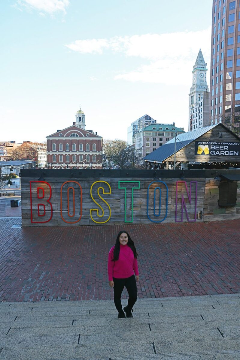 Lily in a pink sweater standing on brick steps in front of a large colorful "BOSTON" sign with rainbow outlined letters, with historic Faneuil Hall, the Custom House Tower clock, and downtown buildings behind her under a blue sky in Boston, Massachusetts